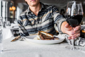 A person in a plaid shirt sits at a table with a plate of steak and mashed potatoes, holding a glass of red wine in a restaurant setting, browsing special dining packages.