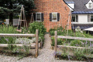 A charming garden with wooden trellises and lush green plants sits in front of a red brick house with black shutters—a perfect Vermont vacation setting. A gravel path winds between raised wooden garden beds.