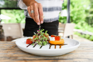 A person’s hand garnishes a plated salad with microgreens at a Casual Fine Dining setting; sliced tomatoes, cheese, and balsamic reduction rest atop a rustic wooden table with a blurred outdoor background.