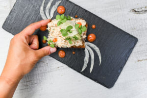 A hand holding a piece of gourmet toast topped with shredded white topping, microgreens, and thin vegetable slices, displayed on a black slate plate with decorative sauce—perfect for a casual fine dining experience.
