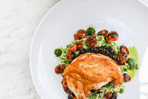 A roasted chicken breast sits on a bed of shredded vegetables, lentils, and roasted cherry tomatoes, garnished with dollops of green herb sauce—perfect for a casual fine dining experience on a white plate.