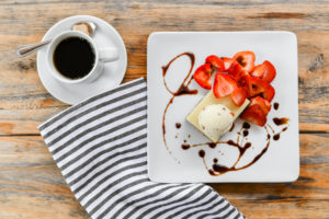 A cup of black coffee with a sugar cube on a saucer sits next to a plate of dessert topped with strawberries, vanilla ice cream, and chocolate drizzle—perfect for a Casual Fine Dining experience on a wooden table with a striped napkin.