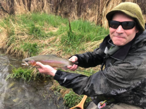 A person wearing sunglasses and a green hat smiles while holding a large rainbow trout by a grassy stream, showing off their fishing success with one of the guided packages.