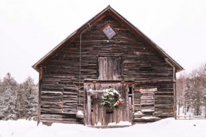 An old wooden barn stands in the snow, decorated with a festive wreath on the door. Snow is falling gently, packages are stacked by the entrance, and evergreen trees are visible in the background.