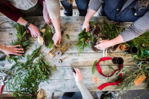 Four people use pinecones, greenery, and ribbon to make holiday wreaths or decorations on a rustic wooden table. Scissors, drinks, scattered foliage, and festive packages are visible in the creative, overhead scene.