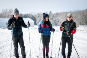 Three people in winter clothing stand on snow with ski poles, smiling and chatting about their ski packages. Snow-covered trees and a white building are in the background on a clear, sunny day.