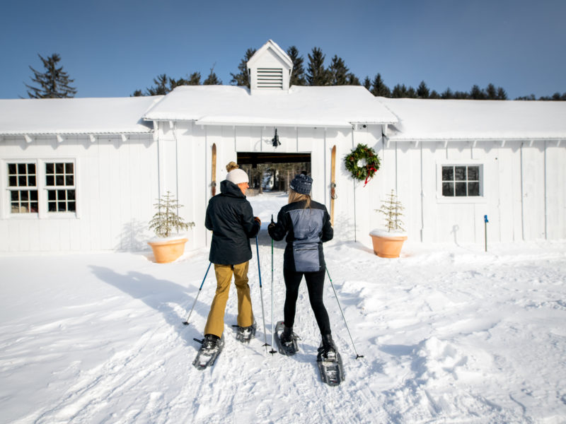 Two people snowshoeing in Stowe VT, dressed in winter clothing, walk toward a white barn adorned with a wreath, surrounded by snow and small potted trees on a sunny winter day.