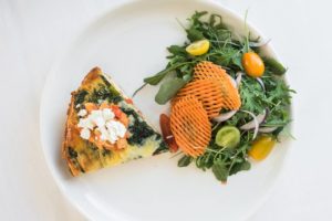 A plate with a slice of vegetable quiche topped with crumbled cheese, served next to a fresh salad of mixed greens, yellow and red tomatoes, red onion, and wavy-cut sweet potato chips.