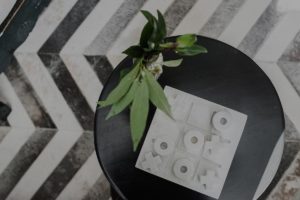 A small black round table with a leafy green plant and a white tic-tac-toe board on top, set against a patterned black-and-white chevron floor—perfect for relaxing during your Vermont Vacation.