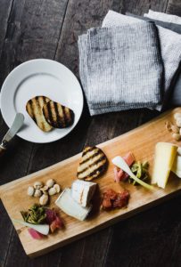 A wooden board with assorted cheeses, grilled bread slices, pickled vegetables, nuts, and preserves sits beside a plate of grilled bread and a butter knife—evoking the rustic charm of Edson Hill - Stowe Vermont Inn.