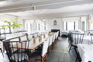 A bright, airy dining room with large windows, a long wooden table set with simple decorations, cushioned chairs, and a stone floor. Light streams in, highlighting the elegant and minimalist decor perfect for casual fine dining.