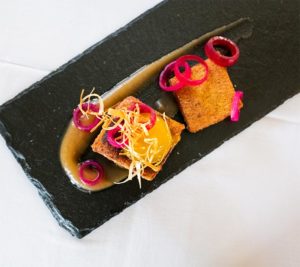 A rectangular black slate plate with two breaded and fried squares, garnished with red onion rings, yellow sauce, and thin vegetable strips, served on a swirl of light brown sauce—perfect for a Casual Fine Dining experience.