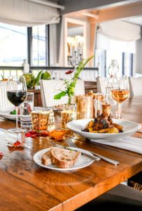 A cozy, elegant dining table set for casual fine dining features wine glasses, water, candles, a flower vase, bread on a plate, and a main dish of meat. Warm sunlight streams in through the windows, creating a welcoming atmosphere.