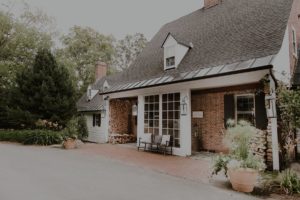 A brick house with a dark gabled roof, white trim, and large windows offers the perfect Vermont vacation vibe. A bench sits by the entrance, with potted plants, stacked firewood, and tall greenery surrounding the building.