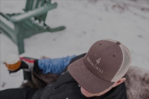 A person wearing a brown Edson Hill cap sits outdoors on a snowy day, enjoying a glass of beer after cross country skiing. A green chair and patches of snow are visible in the background.