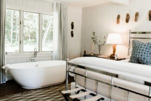 Bright bedroom at Edson Hill - Stowe Vermont Inn features a freestanding white bathtub by a large window, a bed with white linens and blue-patterned pillows, a cozy lamp on a nightstand, and wall art in modern rustic style.