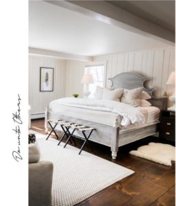 A bright, cozy bedroom with wood-paneled walls, a large bed with white bedding, and wooden floors captures the charm of Vermont luxury hotel accommodations. Two bedside tables with lamps and small benches complete this inviting retreat.