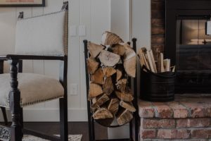 A metal firewood rack holding stacked logs sits next to a black fireplace and a brick hearth, evoking the cozy ambiance of Vermont luxury hotel accommodations. A light upholstered chair and a black container with kindling complete the inviting scene.