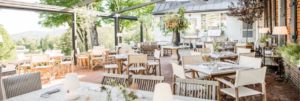Outdoor restaurant patio with white and wooden tables and chairs, decorated with flowers and lamps. Surrounded by greenery and trees, the space is bright and inviting—contact Edson Hill for more information.
