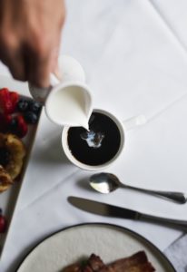 A hand pours cream into a cup of black coffee on a white table. Nearby are a fork, spoon, and plates with assorted breakfast foods, including berries—capturing the cozy essence of breakfast in Stowe, VT.