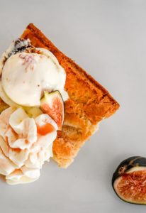 A slice of cake topped with a scoop of ice cream, whipped cream, and a fig wedge sits on a white plate, with a halved fig nearby—a delightful treat often enjoyed during Tavern Nights at Edson Hill.
