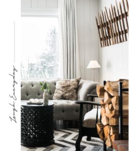 A cozy living room with a tufted sofa, patterned pillow, small round side table, lamp, stacked firewood, and rustic wooden stick wall art; large window with light curtains evokes Vermont Vacation charm. Laugh Everyday text on the left side.
