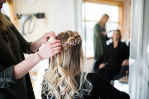 A hairstylist decorates a womans long, wavy blonde hair with a floral hairpiece in a bright salon, preparing her for weddings, while another person sits in the background reflected in a mirror.