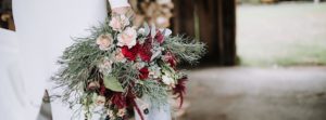 A person in white holds a large, rustic wedding bouquet with pink roses, deep red flowers, and greenery, standing near a wooden structure with an open doorway to the outdoors.