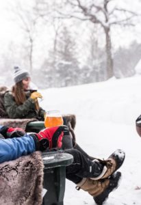 Two people sit outdoors in the snow, bundled in winter clothing after a day of cross country skiing, drinking from glasses. Snow falls gently around them, blankets cover their laps for warmth, and a glass of beer is in the foreground.