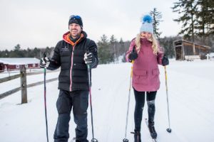 A man and a woman are cross-country skiing on a snowy trail, smiling and dressed in winter clothing, with trees and wooden buildings in the background, enjoying the thrill of cross country skiing together.