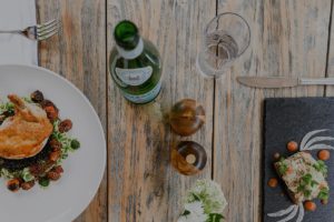 A rustic wooden table set with a plate of chicken and vegetables, a slice of bread with garnish on a slate, a bottle of sparkling water, and elegant utensils—perfect for savoring curated meal packages beside a small vase with white flowers.