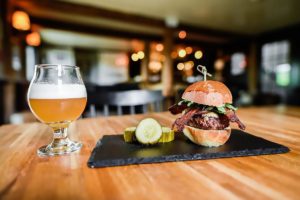 A glass of light beer and a gourmet burger with bacon, lettuce, and a brioche bun, served on a slate plate with pickle slices—perfect for guests enjoying our meal packages in a cozy, warmly-lit restaurant.