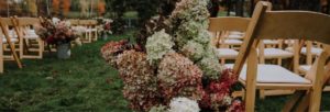 Rows of wooden chairs set up outdoors on grass, with large arrangements of pink, white, and green hydrangeas lining the aisle; perfect for outdoor weddings amid trees with vibrant autumn foliage in the background.