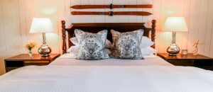 A neatly made bed with patterned pillows, two bedside tables each with a lamp, a small vase of flowers, and a decorative figure. Vintage wooden skis are mounted on a white paneled wall above the headboard, evoking classic Edson Hill charm.