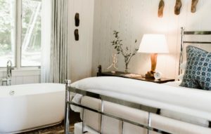 A cozy bedroom features a white freestanding bathtub by a window, a bed with white linens and blue patterned pillows, and a wooden nightstand with a lamp, small plant, and decor. Packages of natural light fill the space.