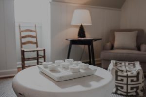 A cozy living room inspired by Edson Hill, featuring a white round table with a modern decorative piece. In the background are a wooden chair, a lamp on a side table, an armchair, and a patterned pouf.