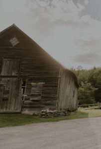 A weathered wooden barn with a slanted roof stands near a gravel road at Edson Hill – Stowe Vermont Inn, surrounded by green grass and trees. Sunlight filters through the clouds, creating a warm, rustic atmosphere.