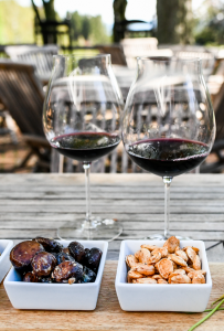 Two glasses of red wine on a wooden table outdoors, with small square dishes from the menu featuring spiced nuts and what appears to be dates or dried fruit. Wooden chairs and trees blur softly in the background.