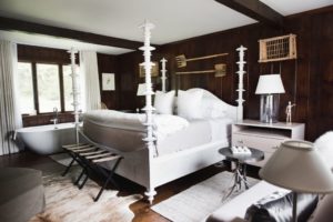 A cozy bedroom with dark wood-paneled walls, a white four-poster bed, a freestanding bathtub by the window, white curtains, a nightstand with a lamp, and eclectic wall decor.