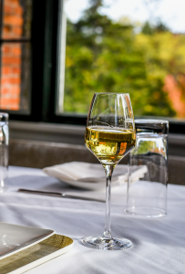A glass of white wine sits on a white tablecloth next to two empty glasses, plates, and napkins, with sunlight streaming in and greenery outside—a perfect setting to explore the menu.
