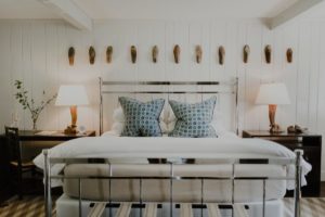 A neatly made bed with a metal frame, two blue-patterned pillows, and white bedding sits between two dark wooden nightstands with matching lamps, evoking the cozy charm of Edson Hill. Wooden shoe molds decorate the white paneled wall above the headboard.