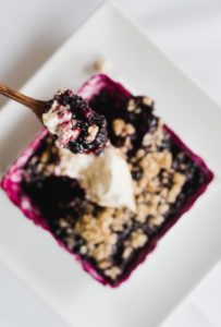 A close-up of a square white dish filled with berry crumble, topped with a dollop of whipped cream. A spoon lifts a portion, revealing juicy berries and crumbly topping—a tempting dessert perfect for any menu.