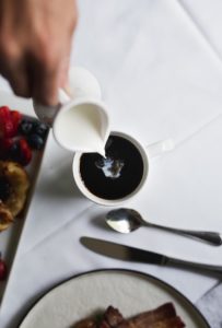 A hand pours cream from a small white pitcher into a cup of black coffee on a white tablecloth, with a plate of fruit and utensils nearby—capturing the cozy charm of breakfast in Stowe, VT.