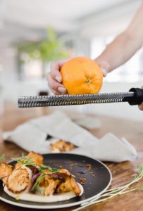 A hand is zesting an orange over a plate of seared scallops and greens on a wooden table, adding fresh aroma. The dish looks ready for the menu, with orange zest falling delicately and a gray napkin nearby against a softly blurred background.