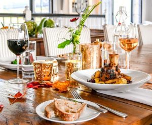 A wooden dining table set for casual fine dining, featuring a plate of bread, cutlery, a dish with meat and vegetables, glasses of red and rosé wine, water, gold candle holders, a small vase with flowers, and autumn leaves as decoration.
