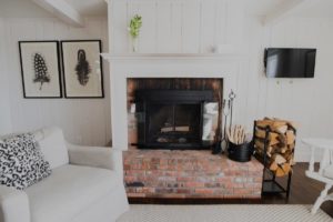 A cozy Edson Hill living room with a brick fireplace, firewood stacked beside it, framed feather artwork on the wall, a sofa with a patterned pillow, and a TV mounted on the wall.