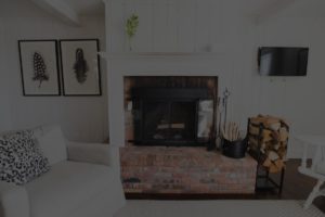 A cozy Edson Hill living room with a brick fireplace, stacked firewood, a white sofa with a patterned pillow, two framed feather prints on the wall, and a small wall-mounted TV.
