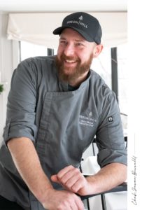 A bearded man in a gray chef coat and black Edson Hill cap smiles as he leans on a table in a bright room with large windows, embodying the warm hospitality of casual fine dining.