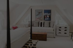 A cozy attic bedroom inspired by Edson Hill Guest Rooms features slanted ceilings, a plush white daybed with gray and white pillows, two white nightstands, a wooden chest, and two framed ski posters above the daybed.
