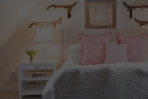 A cozy Edson Hill Guest Room features a striped headboard, pink-patterned pillows, a white nightstand with a lamp and flowers, and decorative antique rifles mounted on the wall.